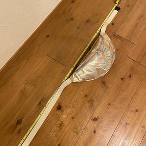 May include: A gold and cream-coloured fanny pack with a floral pattern and a cream-coloured adjustable strap. The fanny pack is measured with a yellow tape measure. The strap is approximately 102 cm long.