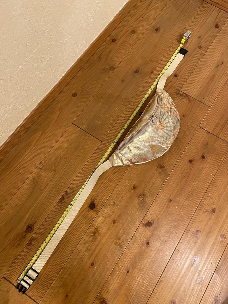 May include: A gold and cream-coloured fanny pack with a floral pattern and a cream-coloured adjustable strap. The fanny pack is measured with a yellow tape measure. The strap is approximately 102 cm long.