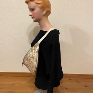 May include: A patterned shoulder bag with a cream strap is displayed on a mannequin. The bag features a gold, green, and orange floral design. The mannequin is wearing a black sweater.