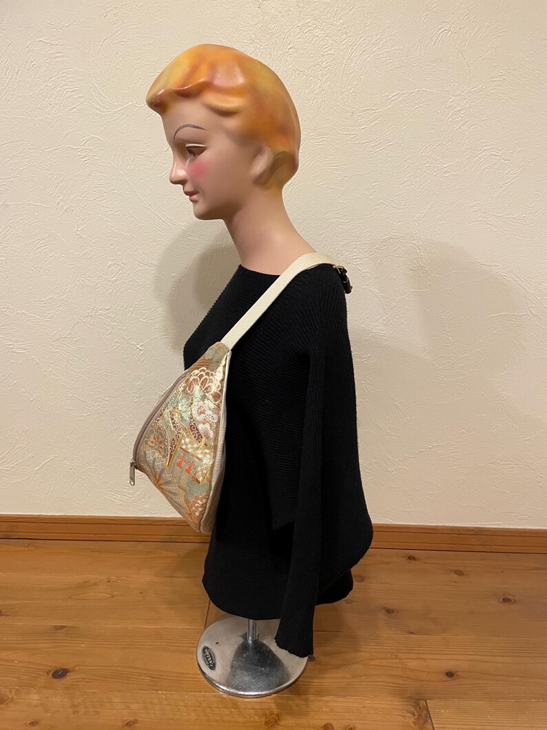 May include: A patterned shoulder bag with a cream strap is displayed on a mannequin. The bag features a gold, green, and orange floral design. The mannequin is wearing a black sweater.