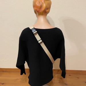 May include: A black, ribbed-knit sweater with a wide neckline and long sleeves is displayed on a mannequin. A beige crossbody strap with a black buckle is worn across the sweater. The mannequin has a tan head and is mounted on a silver stand.