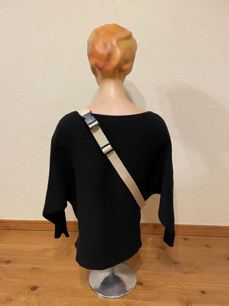May include: A black, ribbed-knit sweater with a wide neckline and long sleeves is displayed on a mannequin. A beige crossbody strap with a black buckle is worn across the sweater. The mannequin has a tan head and is mounted on a silver stand.