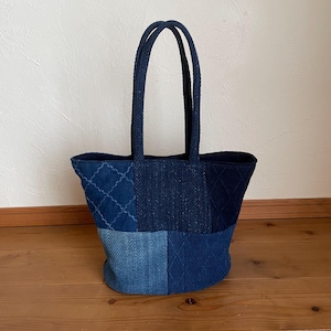 May include: A patchwork tote bag constructed from various shades of blue fabric. The bag features a combination of solid denim-like textures and quilted sections with a diamond-stitched pattern. It includes two matching blue fabric handles.