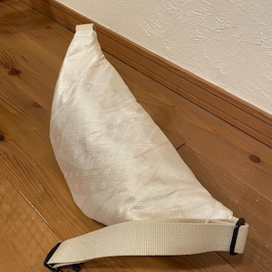 May include: Cream-coloured fanny pack with a geometric diamond pattern. The bag has a long, adjustable strap with a black buckle. The bag is made of a textured fabric and is lying on a wooden surface.