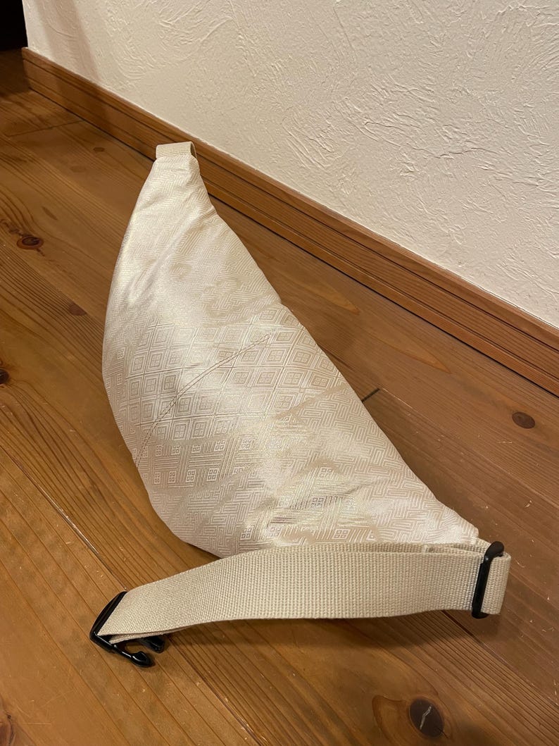 May include: Cream-coloured fanny pack with a geometric diamond pattern. The bag has a long, adjustable strap with a black buckle. The bag is made of a textured fabric and is lying on a wooden surface.