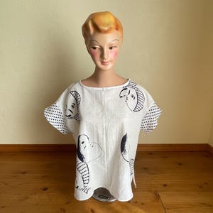 May include: A white short-sleeved top featuring a black graphic print of stylised faces and a polka dot pattern on the sleeves. The garment has a round neckline and a loose, boxy fit, displayed on a mannequin head and shoulders.