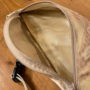 May include: A beige fanny pack with a floral pattern and a black buckle. The interior lining is a lighter beige with a geometric design. The zipper is a matching beige colour.