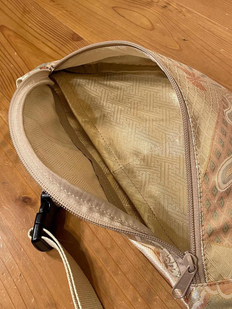 May include: A beige fanny pack with a floral pattern and a black buckle. The interior lining is a lighter beige with a geometric design. The zipper is a matching beige colour.