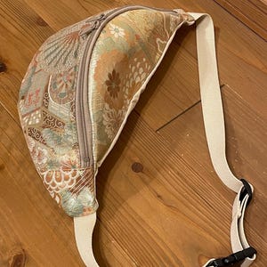 May include: A patterned bum bag with a beige strap and zip. The bag features a floral and geometric design in gold, green, and brown. The strap has a black plastic buckle. The bag is made of a textured fabric.