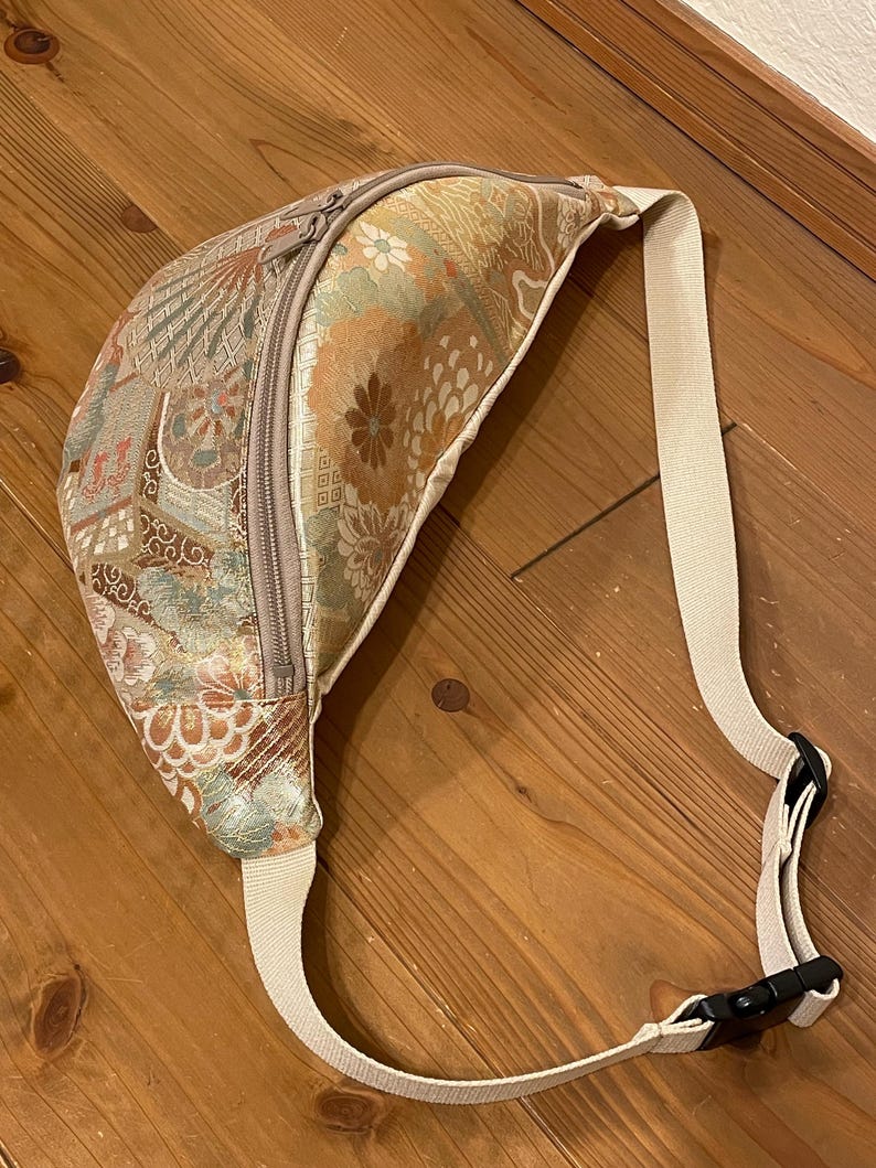 May include: A patterned bum bag with a beige strap and zip. The bag features a floral and geometric design in gold, green, and brown. The strap has a black plastic buckle. The bag is made of a textured fabric.