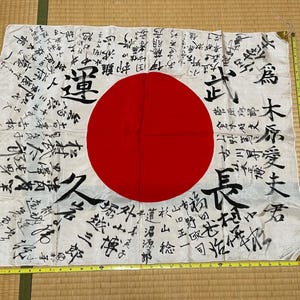 以下が含まれることがあります： 中央に大きな赤い円が描かれた白い長方形の旗で、黒い日本語の書道が全体に書かれています。下端と右端に沿って黄色いメジャーが置かれており、旗の幅が約89cmであることを示しています。