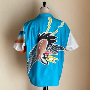 May include: A blue short-sleeved shirt with a white and orange striped pattern on the sleeves. The shirt features a large, colourful graphic of a crane with a red beak and yellow accents.