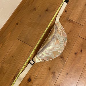 May include: A beige fanny pack with a decorative pattern, a zipper closure, and an adjustable strap. The fanny pack is approximately 30 cm long. A yellow measuring tape is next to the fanny pack.