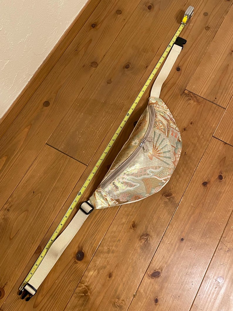 May include: A beige fanny pack with a decorative pattern, a zipper closure, and an adjustable strap. The fanny pack is approximately 30 cm long. A yellow measuring tape is next to the fanny pack.
