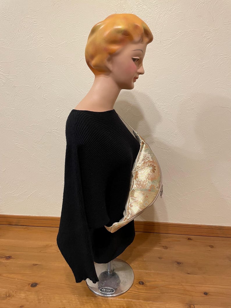 May include: A black, off-the-shoulder sweater on a mannequin. A gold and cream-coloured floral patterned bag is draped over the mannequin's shoulder. The mannequin has a vintage look with blonde hair and a pale complexion.