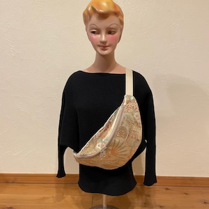 May include: A beige and gold fanny pack with a floral pattern and a cream-coloured strap. The bag is displayed on a mannequin wearing a black jumper. The fanny pack has a zip closure and a curved shape.