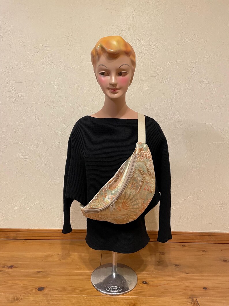 May include: A beige and gold fanny pack with a floral pattern and a cream-coloured strap. The bag is displayed on a mannequin wearing a black jumper. The fanny pack has a zip closure and a curved shape.