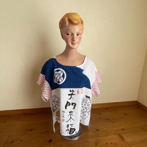 May include: A short-sleeved top featuring a white base with a blue upper section, red and white polka dot ruffled sleeves, and black Japanese calligraphy characters alongside illustrations of figures on the front and a circular emblem.