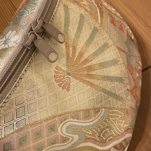 May include: Close-up of a beige fanny pack with a colourful, patterned design. The pattern includes fan-like shapes in orange, blue, and pink. The bag has a beige zip with two pulls. The fanny pack is made of a woven fabric.