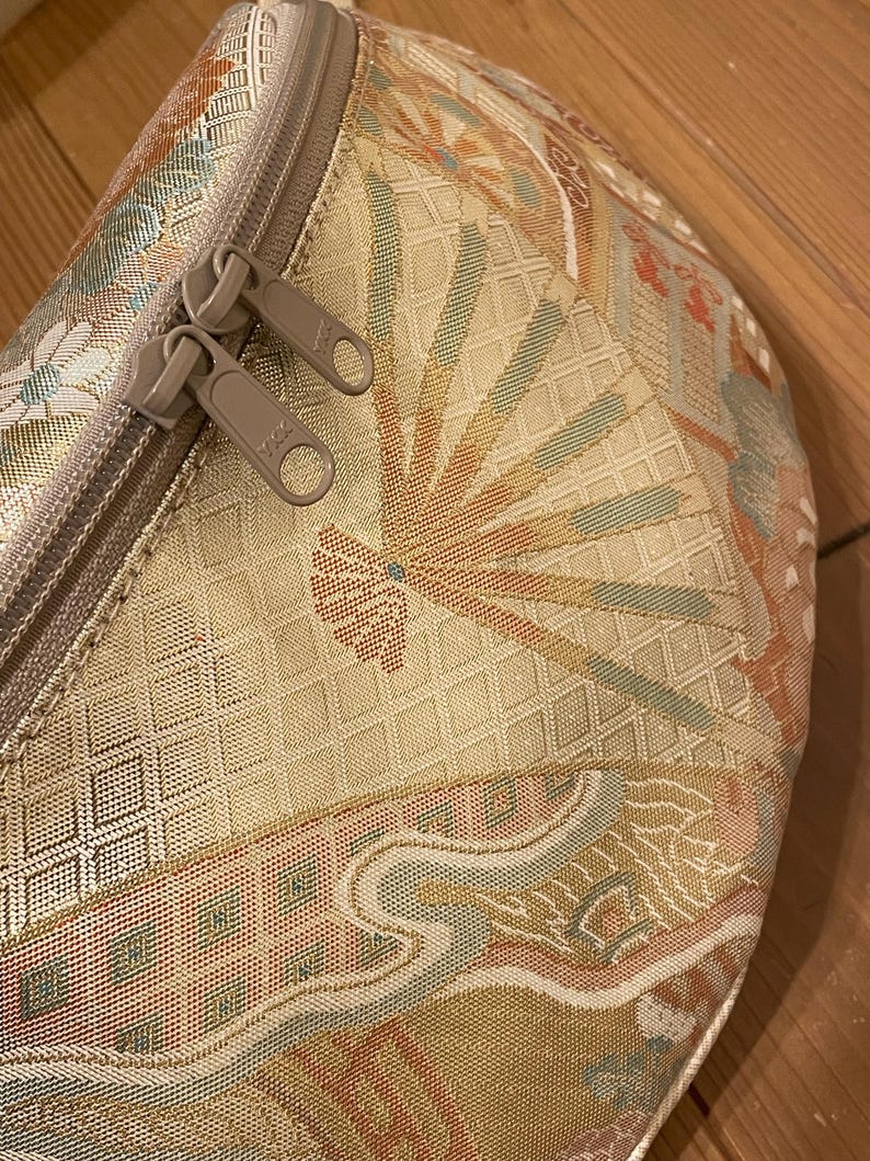 May include: Close-up of a beige fanny pack with a colourful, patterned design. The pattern includes fan-like shapes in orange, blue, and pink. The bag has a beige zip with two pulls. The fanny pack is made of a woven fabric.