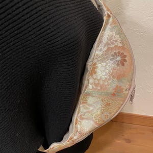 May include: A beige and floral patterned crossbody bag with a zip closure and a thin strap. The bag's interior features a detailed floral design in shades of peach, gold, and green. The bag is shown against a black background.