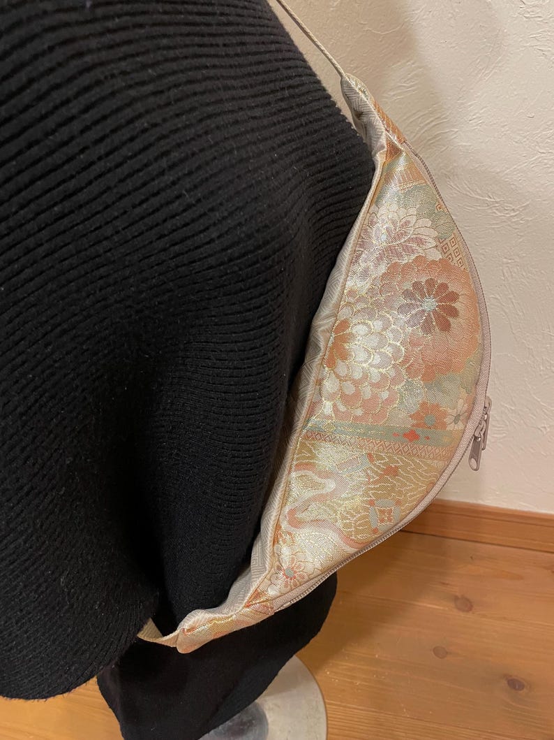 May include: A beige and floral patterned crossbody bag with a zip closure and a thin strap. The bag's interior features a detailed floral design in shades of peach, gold, and green. The bag is shown against a black background.