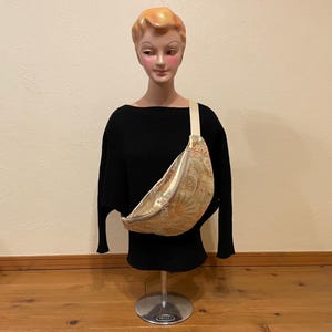 May include: A gold and green patterned fanny pack with a cream-coloured strap and a silver zip. The bag is worn across the body on a black top. The fanny pack has a decorative design.