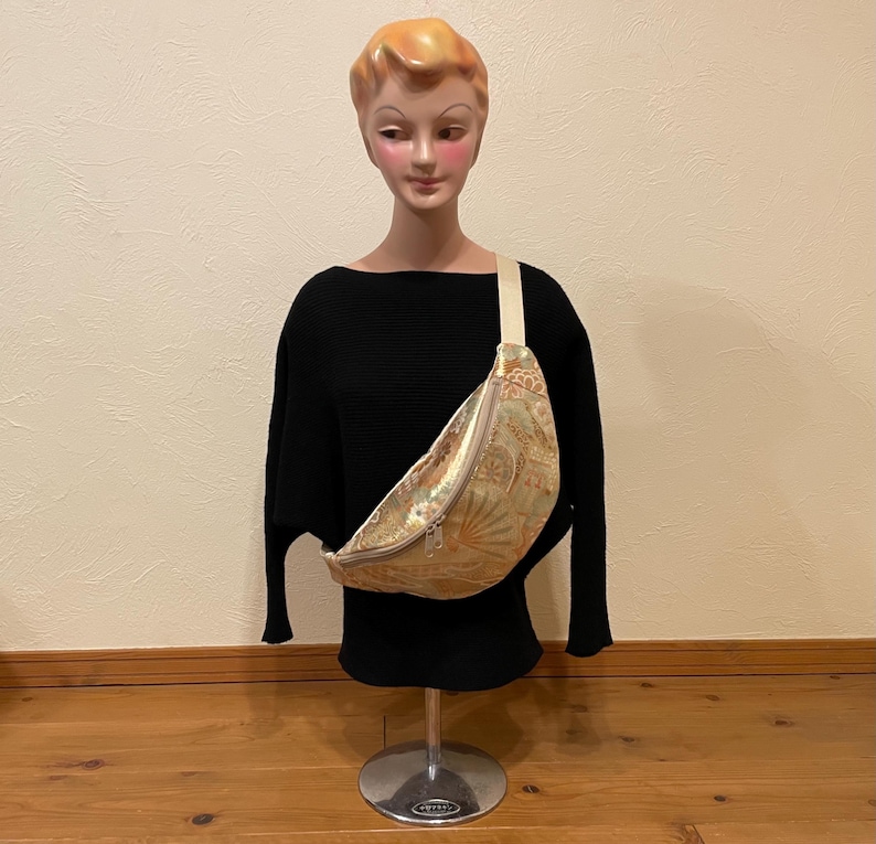 May include: A gold and green patterned fanny pack with a cream-coloured strap and a silver zip. The bag is worn across the body on a black top. The fanny pack has a decorative design.