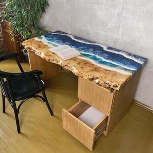 May include: A wooden desk with a unique epoxy resin top, featuring a beach scene with blue and white waves. The desk has a natural wood edge, a drawer, and a black chair. A book rests on the desk.