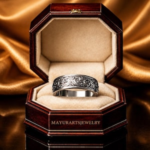 May include: A silver ring with an ornate, swirling pattern sits inside a dark wooden octagonal jewelry box. The box is lined with beige fabric and rests on a reflective surface. The text "MAYURARTSJEWELRY" is visible on the box.