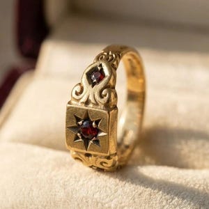 May include: A gold ring with a rectangular face featuring a starburst design with a red gemstone at its centre. The ring has ornate detailing and additional red gemstones. The ring is set against a soft, cream-coloured background.