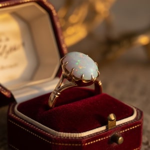 Op de afbeelding: Een gouden ring met een grote, ovale opaal edelsteen. De opaal heeft een melkachtige witte basis met groene en oranje flitsen. De ring is gezet in een klauwachtige zetting en wordt gepresenteerd in een rode fluwelen ringdoos.