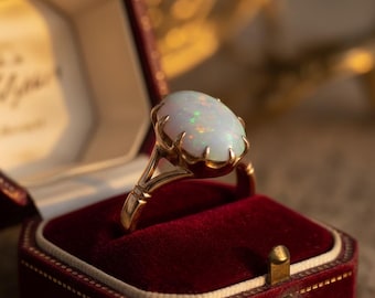 May include: A gold ring with a large, oval opal gemstone. The opal has a milky white base with flashes of green and orange. The ring is set in a claw-like setting and is displayed in a red velvet ring box.