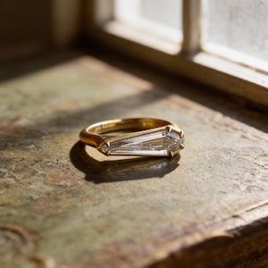 May include: A gold ring with a rectangular, clear gemstone. The ring is set on a textured surface, with light and shadow creating a warm, inviting ambiance. The ring's design is modern and elegant.