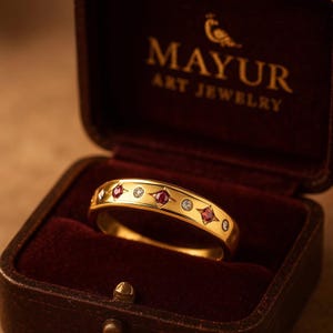 May include: A gold ring with a repeating pattern of red and clear gemstones, displayed in a dark red jewelry box. The ring features the text "MAYUR ART JEWELRY".