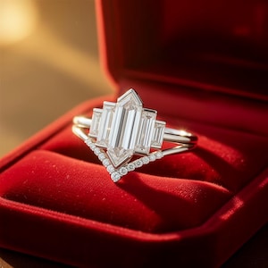 Puede incluir: Un anillo de compromiso plateado con una gran piedra preciosa rectangular y transparente en el centro, flanqueada por piedras rectangulares más pequeñas. El anillo está en una caja de terciopelo rojo, con una banda curva más pequeña debajo, adornada con pequeñas piedras preciosas redondas.