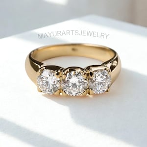 Könnte beinhalten: Ein goldener Ring mit drei runden, klaren Edelsteinen. Der Ring hat ein dickes Band und eine polierte Oberfläche. Der Text "MAYURARTSJEWELRY" ist auf dem Ring sichtbar.