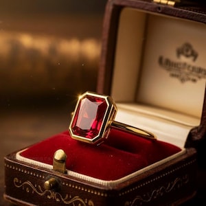Può includere: Un anello d'oro con una grande gemma rossa rettangolare, incastonata in una cornice d'oro. L'anello è posto in un portagioie in pelle marrone foderato di velluto rosso con dettagli dorati e una chiusura decorativa.