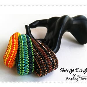 Puede incluir: Cuatro pulseras de cuentas coloridas apiladas en un expositor negro con forma de mano. Las pulseras están hechas con cuentas pequeñas en tonos naranja, amarillo, verde, azul, marrón y negro. El texto "Shanga Bangles: Collin Beading Tutorial" es visible debajo de las pulseras.