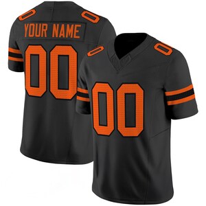Puede incluir: Camiseta de fútbol negra con detalles naranjas, incluido el número "00" en la parte delantera y trasera. La camiseta tiene el texto "YOUR NAME" en la parte superior de la espalda. Las mangas tienen rayas naranjas.