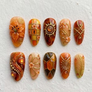 May include: A set of ten press-on nails featuring warm orange, amber, and gold tones. Designs include 3D floral accents, geometric grids with silhouettes, sun motifs, swirling gold lines, lanterns, and intricate leaf patterns with metallic gold details.
