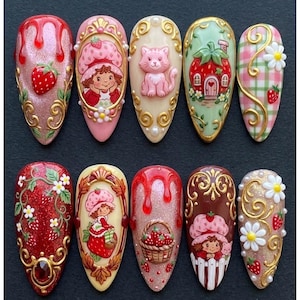 May include: A set of ten almond-shaped press-on nails featuring intricate 3D designs including strawberry motifs, a character in a strawberry hat, a pink cat, a strawberry-shaped house, a basket of strawberries, and gold filigree accents.