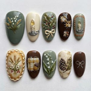 Op de afbeelding: Een set van tien kunstnagels met gedetailleerde 3D-ontwerpen, waaronder lelietjes-van-dalen, kaarsen, dennenappels, eikels en strikken. Het kleurenpalet bestaat uit saliegroen, crème en donkerbruin met verfijnde, reliëfachtige texturen.