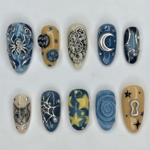May include: A set of ten press-on nails featuring various designs including a silver spider, a clock face with a key, a crescent moon, a cat face, yellow stars, a blue swirl, and a keyhole. The nails are decorated with metallic silver accents.