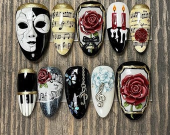 May include: A collection of press-on nails with various designs. The designs include a black and white mask, musical notes, red roses, candles, and a piano keyboard. Some nails have gold accents and glitter.