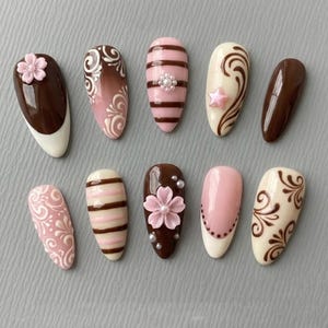May include: A collection of ten artificial nails with various designs. The nails feature a range of colors, including brown, pink, and white, with decorative elements such as floral accents, stripes, and swirls. Some nails have a glossy finish, while others have a matte appearance.