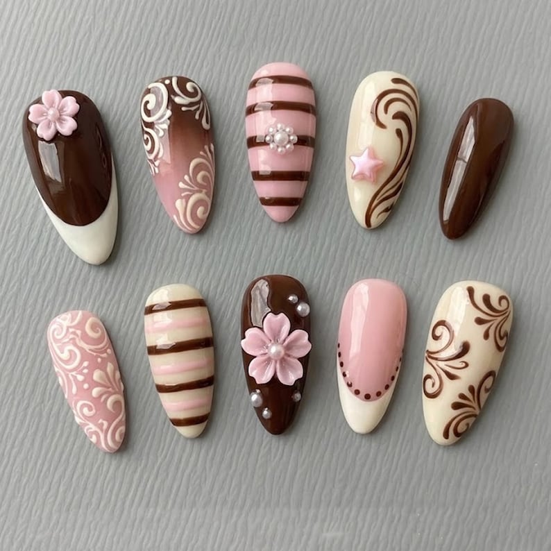 May include: A collection of ten artificial nails with various designs. The nails feature a range of colors, including brown, pink, and white, with decorative elements such as floral accents, stripes, and swirls. Some nails have a glossy finish, while others have a matte appearance.