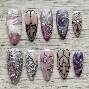 May include: A set of ten artificial nails in shades of pink, purple, and silver. The nails are decorated with intricate designs, including swirls, crosses, and a phoenix. Embellishments include pearls, stars, and metallic details.