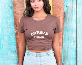May include: A brown crop top with the text "CORGIS 2025" in white. The model is wearing light blue denim shorts. The background is a wooden wall with blue, yellow, and brown paint.