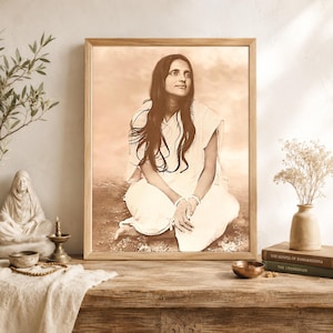 May include: Sepia-toned framed portrait of a woman in white clothing, seated with hands clasped. The image is displayed on a wooden table with a small brass bowl, books, and a vase with dried flowers. A statue and candle holder are also visible.
